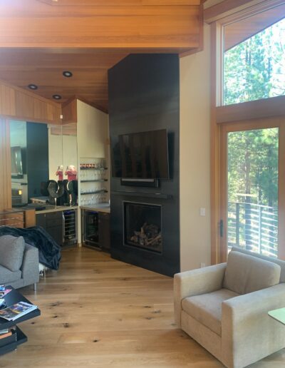 Modern living room with a fireplace, hardwood floors, and large windows with a forest view.