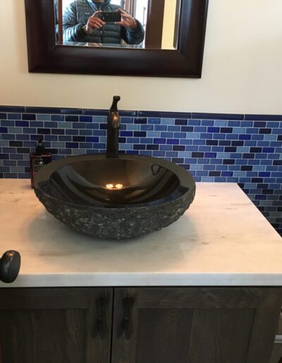 Person taking a photo of a bathroom with a stone vessel sink and blue mosaic tile backsplash.