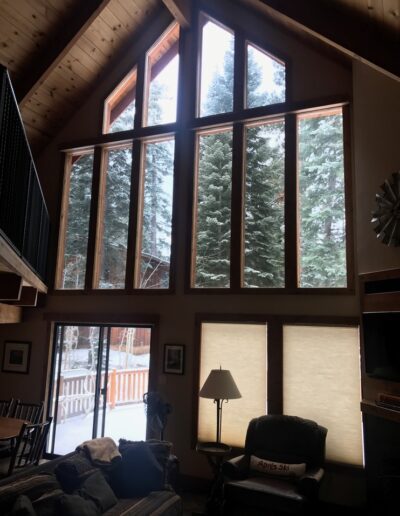 A cozy living room interior with a high vaulted ceiling, large windows revealing snow-covered trees, and comfortable furnishings.