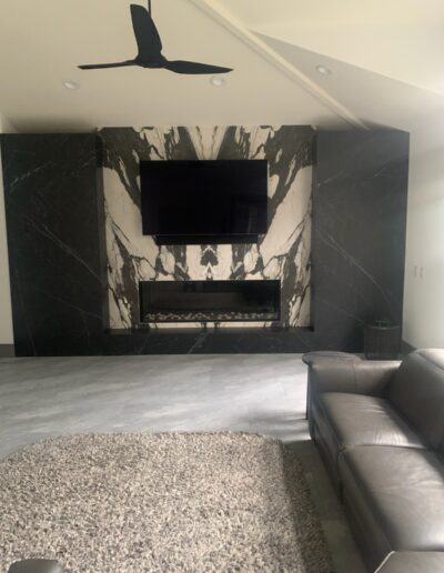 Modern living room with a wall-mounted television above a fireplace, surrounded by dark marble, complemented by minimalist furnishings and a ceiling fan.