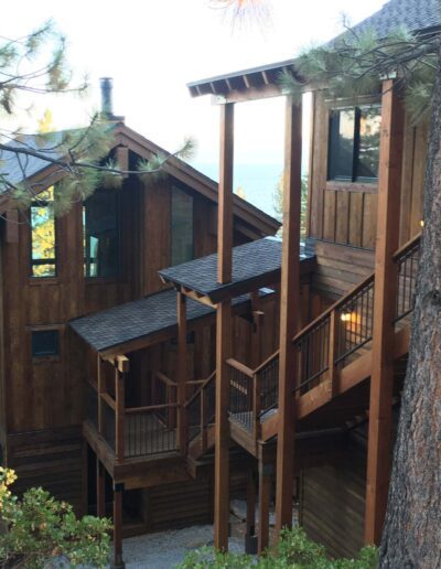 Wooden cabin with exterior staircases surrounded by trees with a view of the water in the distance.